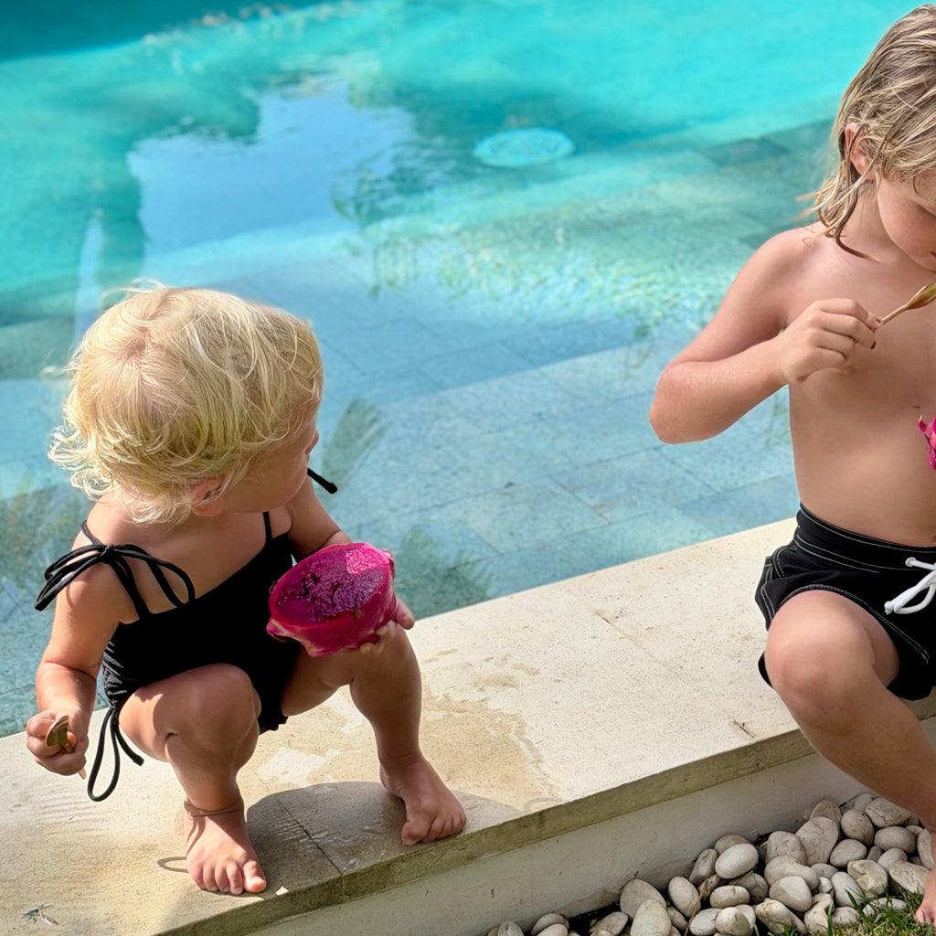 Two children by a pool, one in a black  swimsuit with pink flower, the other in black board shorts.