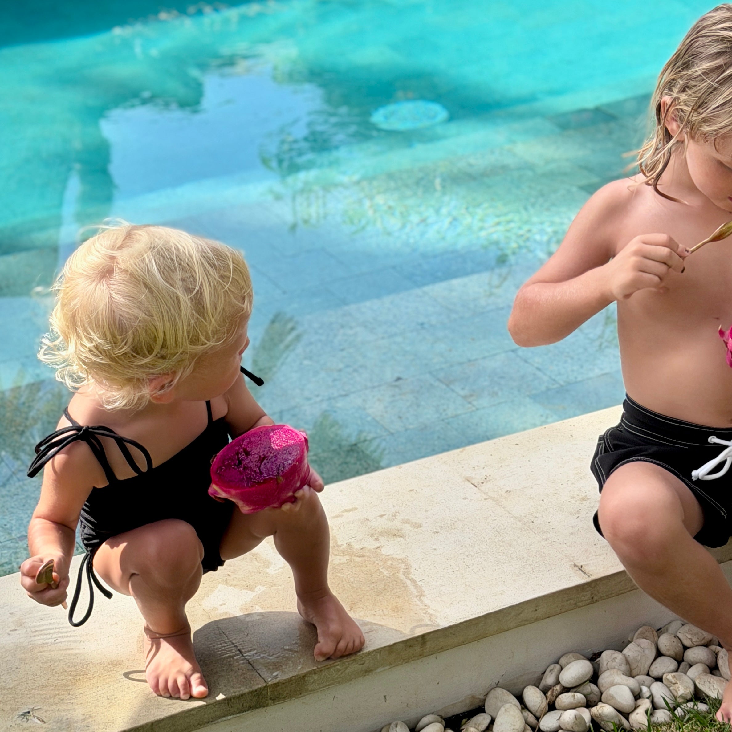 Two children by a pool, one in a black  swimsuit with pink flower, the other in black board shorts.